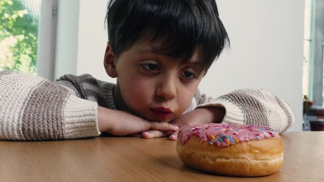 Boy stares longingly at a pink frosted donut with sprinkles on a wooden table, thoughtful and curious expression in a brightly lit room