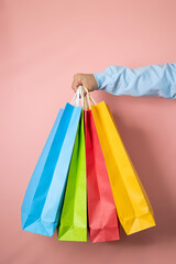 Male hand in blue diver peeking out multicolored shopping bags on a pink background, consumerism...