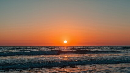 Fototapeta premium Summer sunset scene over tranquil waters featuring soft blue hues and a bold pumpkin-colored sky. Peaceful waves flow from the horizon.