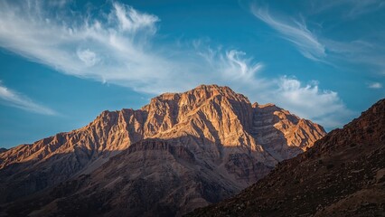 Obraz premium Mountain landscape under blue sky with clouds showcasing rugged peaks and rocky terrain bathed in warm sunlight during daytime.
