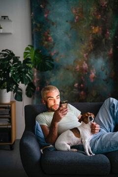 A man with blonde hair sits on a sofa, engrossed in his smartphone, its light illuminating his face. His small dog rests beside him, looking up, as he pets it.