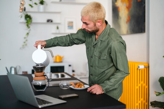 A young man with blonde hair prepares pour-over coffee in his modern kitchen. He pours hot water into the coffee dripper, with a laptop and breakfast pastries nearby.