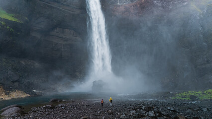 Adventurers stand in awe at the base of a powerful Haifoss waterfall in Iceland, surrounded by...