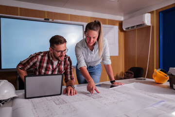 Two engineers are examining blueprints on a construction site, using a laptop and tablet for their analysis, working together to ensure the project's success within their temporary office