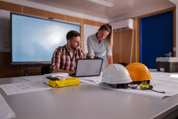Two construction engineers are working on a building project in their office, using a laptop and...