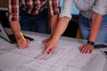 Two architects are bending over a table and reviewing a blueprint of a building project, pointing...