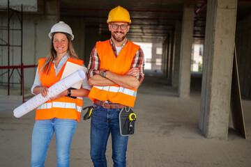 Two smiling architects standing in a construction site, wearing safety vests and helmets while...