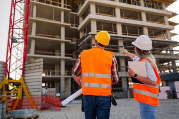 Two construction workers, a man holding blueprints and a woman with a tablet, are discussing the...