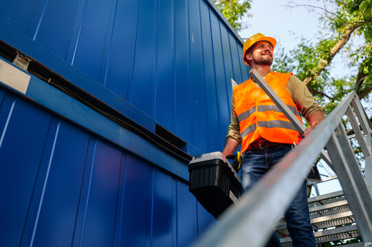 Construction worker wearing safety vest and helmet climbing metal stairs with toolbox and level, working on a construction site next to a blue container