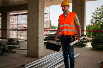 Construction worker wearing safety vest and helmet carrying toolbox and spirit level walking on...
