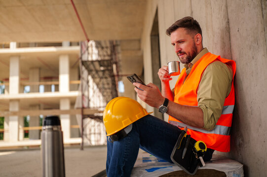 Construction engineer wearing safety vest and hard hat drinking coffee and using smartphone during break on construction site, building and scaffolding in background