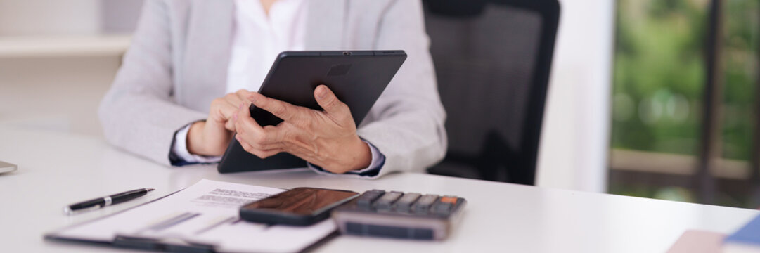 Adult Asian professional uses digital tablet for financial analysis calculating business strategy and data reporting in modern office workspace - Powered by Adobe