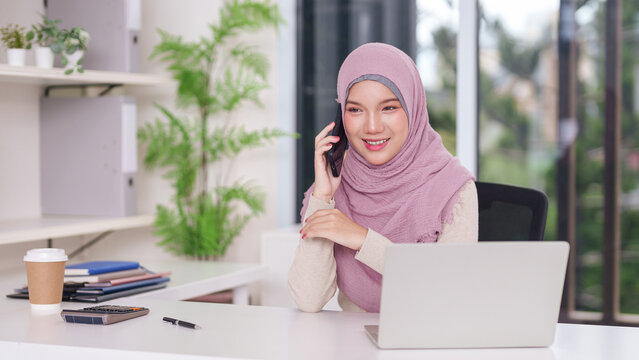 Asian adult Muslim businesswoman actively communicating phone laptop modern office entrepreneur work success digital connection professional workspace productive environment