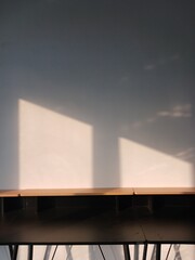 Abstract shot of sunlight streaming across a grey and white wall casting shadows near a wooden frame and dark furniture