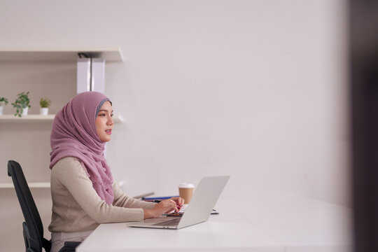 Adult Asian Muslim woman works on laptop computer calculating finances at modern office desk professional business productivity