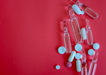 A composition of clear medical ampoules and various colored pills scattered across a vibrant red...