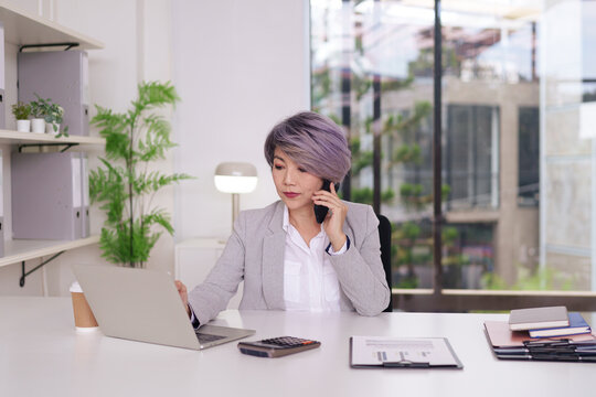 Asian adult female executive professional working modern office multitasking laptop phone call corporate business manager entrepreneur communication technology workspace efficiency - Powered by Adobe
