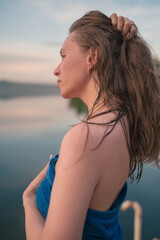 A young woman with wet hair wrapped in a blue towel stands near a calm lake at sunset, gazing thoughtfully into the distance.