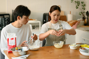 Happy woman cracking an egg while her partner whisks flour to prepare cookie dough