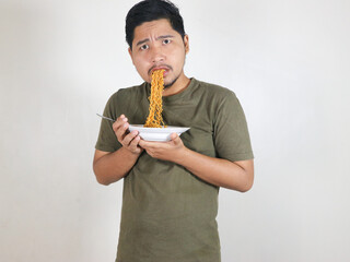 Asian handsome man eating noodles with a fork and showing a shocked and stunned expression. eating...