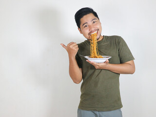 Handsome Asian man enjoying noodles, with the noodles still in his mouth, and pointing to the side. eating lunch concept. isolated white background