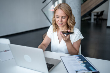 Businesswoman recording voice message on smartphone while working on laptop