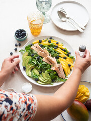 Woman adding salt to fresh healthy salad with chicken, mango, avocado and blueberries