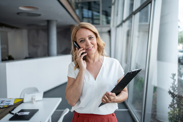 Businesswoman talking on phone and holding clipboard in modern office