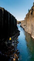 Two adventurers stand along the rocky banks of Studlagil Canyon, marveling at the towering basalt...