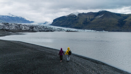 Two adventurous hikers walk along the black sand shores near Skaftafell Glacier, surrounded by...