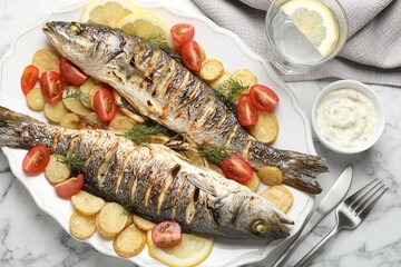 Tasty roasted seabass fish served with potatoes, tomatoes, sauce and lemon on white marble table, flat lay. Homemade seafood dish