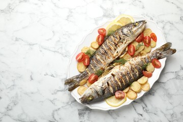 Tasty roasted seabass fish served with potatoes, tomatoes and lemon on white marble table, top view and space for text. Homemade seafood dish