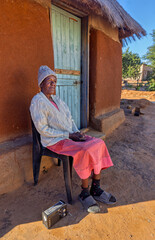 Africa village, single old african woman listening to portable radio outdoors, sited on a chair in front of the thatched roof mud house