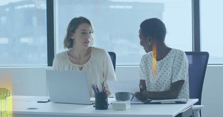 Sitting colleagues in blouses collaborating at office desk by window, with laptop and documents