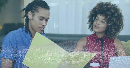 Coworkers reviewing yellow folder on gray sofa in office lounge, with silver laptop and coffee cup
