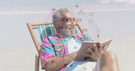 Reclining senior man holding paperback on sandy beach near shoreline, with digital icons