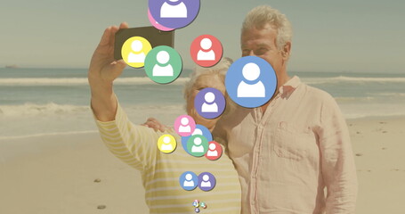 Taking selfie older couple standing on sandy beach, with smartphone and floating social media icons
