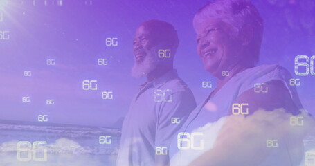 Standing mature couple gazing toward horizon on sandy beach, with floating 6G digital symbols