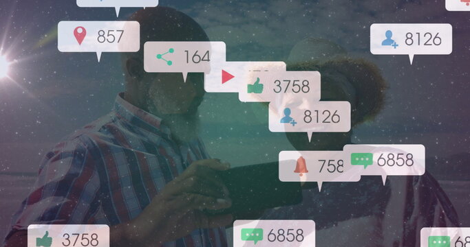 Mature couple in plaid shirt, straw hat, sunglasses holding tablet at beach, showing notifications