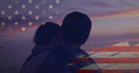 Hugging adult couple standing at shoreline under pastel sky, with ocean water and flag overlay