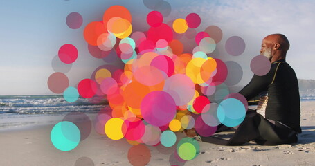 Sitting senior man wearing wetsuit gazing at ocean on sandy beach, with colorful bokeh, copy space
