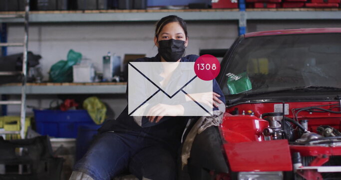 Leaning female mechanic wearing coveralls and mask on red car chassis in workshop, with mail icon