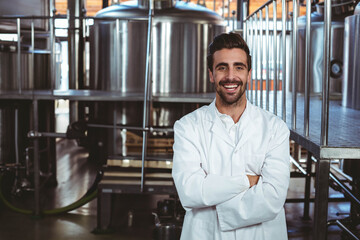 Male brewery technician wearing lab coat crossing arms on brewery floor near fermentation tanks