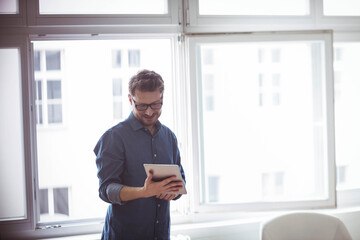 Fototapeta premium Man wearing glasses and blue button-up shirt standing by window using tablet in office, copy space
