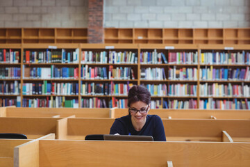 Woman wearing glasses working at study carrel in academic library with laptop, notebook and pen