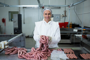 Mature adult male worker wearing hairnet, gloves handling sausages on conveyor belt at meat plant