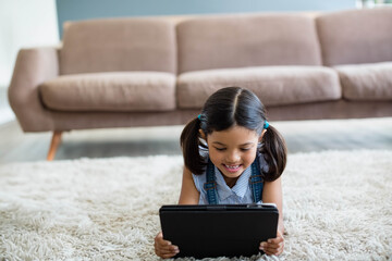 Girl holding black tablet lying on white shag carpet in living room with brown sofa