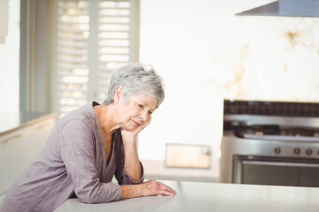 Senior woman leaning on stone countertop near shuttered window, gazing downward by steel stove
