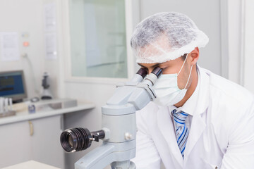 Lab technician peering through light microscope on bench, with computer monitor and pipette stand