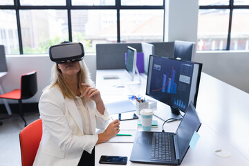 VR headset resting on office desk with dual monitors showing graphs and coffee cup near window